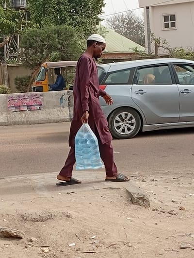 A male subject is observed walking from right to left across the lower-center of the frame, on an unpaved shoulder adjacent to a paved road in Darmanawa, Nigeria. He wears a maroon long-sleeved tunic (kaftan) and matching trousers, with dark-colored open-toed slide sandals on his feet. A white skullcap (kufi) is worn on his head. He carries a transparent plastic bag containing multiple individual sachets of clear liquid, likely water, in his right hand. His gaze is directed downwards and slightly forward.
In the mid-ground, immediately behind the subject, a silver station wagon or minivan is partially visible, moving in the same direction. A person, possibly female, with a light-colored head covering, is visible through the rear passenger window of the silver vehicle. Further left in the mid-ground, a yellow three-wheeled motorized rickshaw (Keke Napep) is partially visible behind a concrete road barrier, with a male driver in a blue shirt visible inside.
The environment includes a dirt shoulder in the foreground with scattered debris and a low concrete curb. Behind the road barrier and vehicles, green trees and bushes are visible, with a light-colored building featuring a single square window in the upper-right background. The sky appears overcast or hazy.