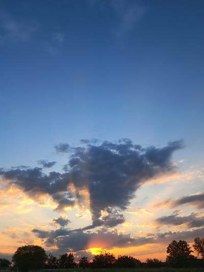 The image depicts a sunset scene over a rural landscape in Volodymyrivske, Ukraine. The sky is dominated by a gradient of blues transitioning into vibrant oranges and yellows near the horizon, indicating the time of day is late afternoon or early evening. Scattered clouds, ranging from dark gray to wispy white, are illuminated by the setting sun, creating dramatic shafts of light that pierce through them. The lower portion of the image shows a silhouette of trees and vegetation against the bright sky, suggesting an outdoor, natural setting. The overall mood is serene and picturesque, characteristic of a peaceful sunset. There is no visible text in the image.