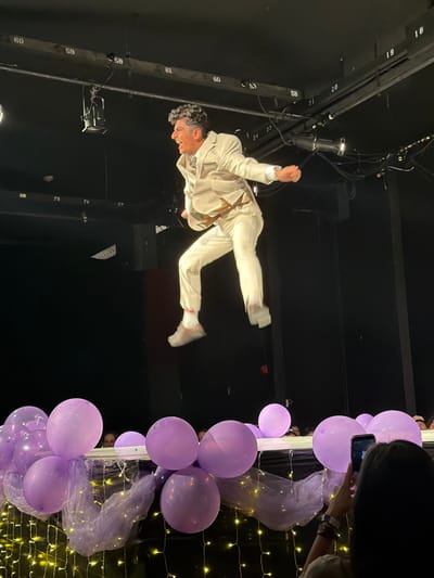 The image captures an indoor stage performance in Palos Verdes Estates, United States. A male individual, appearing mid-air, is the central subject, dressed in a light-colored suit with a light green tie, white socks, and light-colored athletic shoes. His dark, curly hair is visible, and his mouth is open in an expression of excitement or exertion, with both hands clenched. Overhead, exposed metal rigging with stage lights and numerical labels, such as "58" and "59", is visible against a dark background. The lower portion of the frame features a decorated barrier adorned with multiple light purple balloons and sheer purple fabric, illuminated by yellow string lights. Partially visible audience members are situated behind this barrier. In the bottom right corner, a person with dark hair, viewed from behind, is holding a smartphone horizontally, appearing to record the performance.
