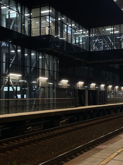 The image depicts a modern train station platform at night in Aprelevka, Russia. The station features a multi-level glass and metal structure with illuminated interior spaces. A wooden platform runs along the train tracks, with railings in place for safety. The train tracks themselves are clearly visible in the foreground, laid with ballast. The overall impression is one of contemporary infrastructure and efficient design, with the artificial lighting creating a stark contrast against the dark night sky. No individuals or specific actions are discernible in the scene.