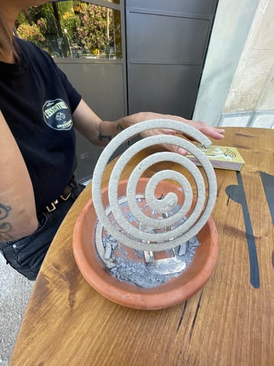 A person wearing a black t-shirt and denim jeans is seated at a wooden table outdoors. The person's left hand is resting on a mosquito coil, which is coiled in a spiral and placed in a terracotta dish filled with ash. To the right of the mosquito coil, a box of matches is visible on the table. In the background, a reflection in a window shows greenery and a building. The overall scene suggests an outdoor setting, possibly a patio or cafe area, in Cucuron, France.