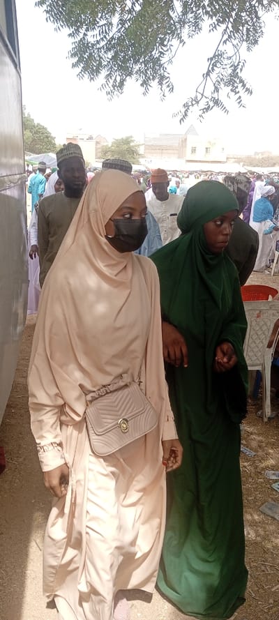 This image depicts a daytime outdoor scene in Sani Mai Nagge, Nigeria, showing two women in the foreground amidst a larger gathering of people. The woman on the left wears a light beige full-length garment and hijab, a black face mask, and carries a beige quilted crossbody bag with a gold clasp. Her left hand is visible at her side, with red nail polish on her fingernails. The woman on the right wears a dark green full-length garment and head covering; her right hand is holding the left forearm of the woman in beige. Both women are walking or standing on unpaved, dusty ground.
In the mid-ground, numerous individuals, primarily men wearing various traditional caps and light-colored clothing, are visible, suggesting a crowd or event. Some women are also present in the background. A large, dark-colored structure, possibly a vehicle or building, is partially visible on the far left. Distant buildings with light-colored walls and flat roofs are present in the background. Overhead, green tree foliage is visible against a bright, overcast sky. Debris, possibly paper currency, is scattered on the ground in the lower right foreground. Partially visible white plastic chairs are on the right edge.