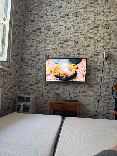 The image depicts an indoor scene, likely a bedroom, with a television mounted on a wall covered in a grey and white patterned wallpaper. The television displays a cooking show featuring a pot of soup with steam rising from it. To the left of the television, a window with old, possibly white-painted wooden panes is visible. Below the television, there is a small wooden nightstand with a drawer. In the foreground, two white mattresses are laid out, suggesting a bed. To the right of the television, an electrical outlet with a plug and wires is mounted on the wall, and a black electrical cord extends downwards. A small black fan is partially visible on the far right. On the wall between the television and the fan, there are several small decorative items or figurines. On the left side of the image, behind the mattresses, is a large object covered in clear plastic sheeting, possibly a piece of furniture or an appliance. The overall impression is of a lived-in, somewhat cluttered domestic space. The location is identified as Tarasovsky, Russia.