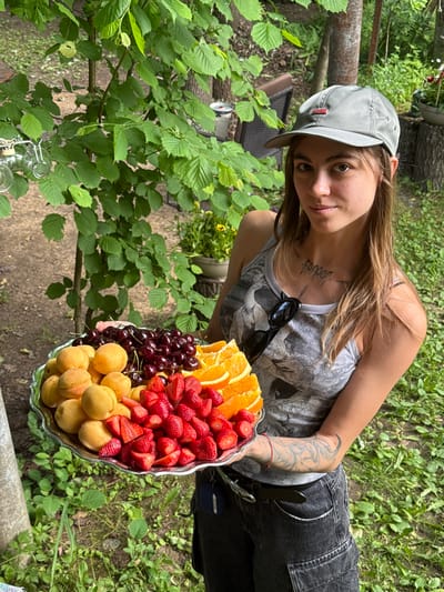 A young woman wearing a gray baseball cap and a gray tank top with a bird print is holding a large silver platter of fruit. The platter contains apricots, cherries, sliced oranges, and strawberries. She has tattoos on her arms and chest. She is standing in an outdoor setting with green trees and foliage in the background. The ground is covered with grass and dirt. The image appears to have been taken during the daytime. The specific location is Буран Снт, Russia.