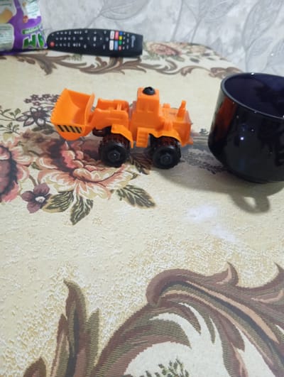 A toy excavator is situated on a patterned fabric surface. To the left of the excavator, a bag of chips is partially visible. Behind the excavator and slightly to the right, a black mug is present. In the background, a remote control is visible. The scene appears to be indoors, possibly on a table or similar surface, with a patterned tablecloth. There are no people depicted in the image. The specific location in Charshanba, Uzbekistan cannot be determined from the visual information provided.