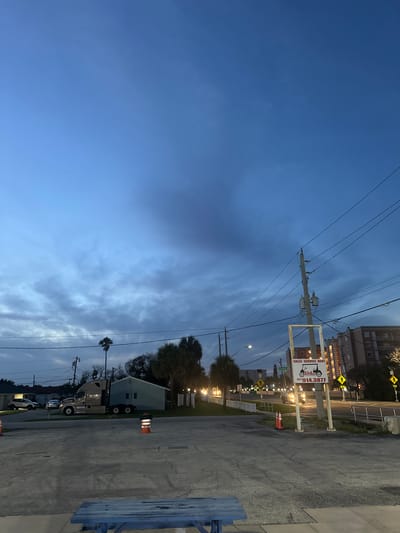 The image depicts an outdoor scene at dusk in Indialantic, Florida, United States of America. The sky is a deep blue with scattered clouds. In the foreground, a weathered blue picnic table is partially visible. The middle ground features a paved area, a light blue building, a large semi-truck, and several orange traffic cones. Palm trees and other vegetation are scattered throughout the scene. In the background, a street with traffic lights and buildings can be seen, including a multi-story building that appears to be an apartment complex. A sign for "Cartbarn" with a phone number and services offered is mounted on a pole. Power lines and utility poles are prominent throughout the image. The overall impression is a commercial area transitioning from day to night.