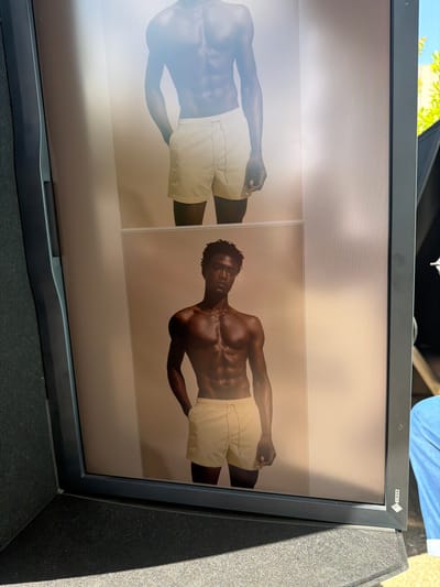 The image shows a monitor screen displaying a picture of a shirtless, muscular man wearing light-colored shorts. The same image is mirrored and appears twice on the screen. The monitor is black and located outdoors. The location in the city of Fibracolor, Spain is not identifiable from the given image.