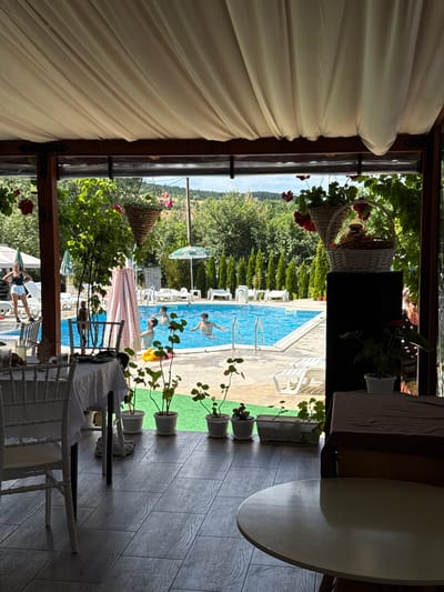 The image depicts an outdoor seating area with a view of a swimming pool, set in Lozno, Bulgaria. In the foreground, there are tables and chairs, one with a white tablecloth and another with a brown cloth. Several potted plants are arranged along the edge of the seating area. Beyond the seating, a blue swimming pool is visible with several people swimming and interacting. Lounge chairs and umbrellas are situated around the pool. The background features lush green trees and a hillside. The overall scene suggests a recreational or dining establishment with a pool.