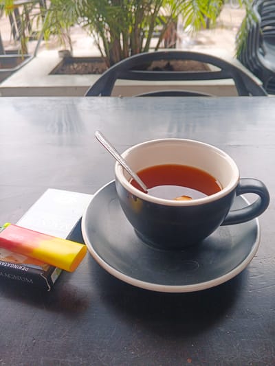 A close-up, eye-level, outdoor shot captures a dark-colored ceramic mug filled with tea and a spoon resting inside. The mug sits on a matching saucer, both placed on a textured, dark tabletop. To the left of the mug are a pack of "MAGNUM FILTER" cigarettes and a brightly colored cigarette lighter, featuring red and yellow hues, with the brand "Cricket" visible on its side. The background is blurred, showing glimpses of greenery, possibly plants and trees, suggesting an outdoor seating area at a cafe or restaurant. The overall lighting is natural, indicating it is daytime. The scene is set in Geulumpangsulu Timu, Indonesia.