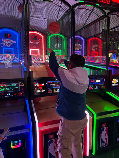 The image shows a person playing an NBA Hoops basketball arcade game. The game has multiple hoops with different team logos: Oklahoma City Thunder, Chicago Bulls, Los Angeles Lakers. The player is shooting a basketball towards the Chicago Bulls hoop, which currently has a score of 21. The score on the Oklahoma City Thunder hoop is 0. The person is wearing a blue and grey sweater and khaki pants. The game features illuminated LED lights and displays the high score as 255. The location of the game is the city of Columbus, United States of America.