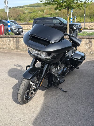 A black Harley-Davidson touring motorcycle is parked on an asphalt surface in Grevenmacher, Luxembourg. The motorcycle is positioned at an angle. Behind the motorcycle is a black SUV, followed by a grey Citroen C3, identified by a license plate beginning with "TR42", parked along the side of the road next to a sidewalk. A tree is located next to the SUV, with street signage visible behind the tree. The environment includes a hillside with trees in the distance. The sky is light, suggesting daylight. The motorcycle is casting a shadow on the asphalt.