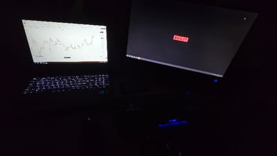 A dark desk setup features two screens. The screen on the left displays a white stock chart, with a keyboard illuminated by white LEDs visible below. To the right, a larger screen showcases a red rectangle with the word "VALVE" on it. Blue lighting subtly illuminates objects on the desk, though most remains obscured by darkness.