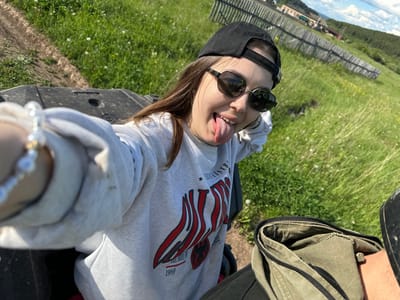 A person, presumably female, is lying on their back in a grassy area. The person is wearing a grey sweatshirt with a red and white design, sunglasses, and a black baseball cap. Their tongue is sticking out. They appear to be taking a selfie with a phone visible in the foreground, near a bracelet. The location is potentially in the city of Пепеляева, Russia. A wooden fence and what seems like a body of water are visible in the distance. Part of an ATV (All-Terrain Vehicle) is visible in the frame. The grass is green and has white flowers.