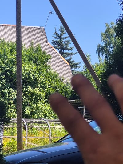 The media file shows an outdoor scene with a person's hand prominently in the foreground, obscuring part of the view. The hand appears to be in focus, while the background elements are slightly blurred. Behind the hand, a tiled roof of a building is visible, along with lush green trees and a bright blue sky. There are also two horizontal, weathered metal bars stretching across the frame, suggesting they are part of some structure or fencing. A metal antenna extends from the roof of the building. In the right side of the image, a dark blue or black reflective surface, possibly a vehicle, is partially visible. The lighting suggests it is daytime, likely sunny, given the bright sky and clear shadows. The overall impression is a snapshot taken outdoors on a clear day, possibly from a low angle looking up, with the photographer's hand partially covering the lens. The location context of Švenčionys, Lithuania, places this scene in an Eastern European setting. No text is visible in the image.