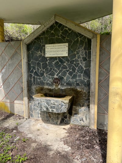 A small water fountain is constructed of stone and concrete. The facade is made of randomly arranged black rocks. A trough-like stone catches the water. A metal spigot in the center of the stone facade is connected to the water supply by a rusty metal chain. A sign to the left of the spigot reads "Cheshmata e postroena 2006g. sus sredstva dareni ot Hyusein Hasan Saroglu ot grad Nikopol." The background is filled with green vegetation and dirt. The scene is located in Nikopol, Bulgaria.