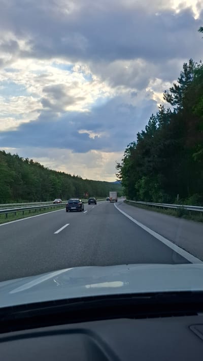 The image shows a view from inside a vehicle driving on a multi-lane highway. The highway is flanked by dense green trees on both sides, with a cloudy sky overhead. Several cars and a truck are visible on the road. The perspective is from the driver's seat, looking forward. The scene appears to be a highway outside a city, given the presence of trees and the open road. There is no specific indication of the location being the city of Gorno Varshilo, Bulgaria.