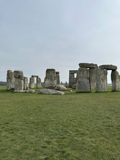 The image displays Stonehenge, an ancient megalithic monument located approximately 2 miles west of Amesbury, United Kingdom. The scene is dominated by a partial circular arrangement of large, weathered sarsen stones, varying in size and orientation. Several stones stand upright, some forming complete trilithons with horizontal lintels across two vertical supports. Other stones are visible lying horizontally on the ground around the central formation. The immediate environment consists of a vast expanse of well-maintained, short green grass. In the background, beyond the stone circle, a flat, open landscape extends to a distant treeline under a light grey, overcast sky. No individuals or active elements are present within the frame.