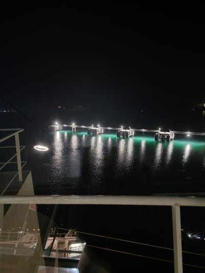 The image shows a nighttime view of the Brindisi port in Italy, specifically looking out towards a pier or terminal extending into the water. The water is illuminated with a bright green glow, likely from underwater lights, and reflects the lights from the pier and distant structures. On the left side of the image, part of a ship or ferry, from which the photo is taken, is visible, including railings and a porthole. In the foreground, the reflection of another ship or structure, with the partially visible text "TRIESTE" in reverse, is seen on a dark, wet surface. The overall atmosphere is dark with scattered artificial lights.
