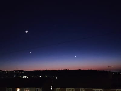 The image depicts a twilight scene in what appears to be Holywell, United Kingdom. The sky transitions from a deep blue at the top to a reddish hue near the horizon. A crescent moon is visible in the upper left quadrant of the sky, with two bright stars or planets positioned below it and closer to the horizon. Power lines run horizontally across the frame. Below the horizon, a silhouette of rolling hills and rooftops is visible, dotted with lights suggesting urban or suburban development. A television antenna is silhouetted against the horizon on the right. The foreground shows the roofs of residential buildings, some with illuminated windows.