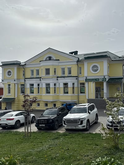 The image shows a three-story yellow and white building with green trim, identified as a Sberbank branch ("СБЕР" is visible on a sign in Cyrillic). Several cars are parked along the curb in front of the building. The license plates of the cars are visible. The license plate of the first car from the bottom is "G399TT 102", the second car is "A515MX 252", the third car is "B405EA 17", and the fourth car is "X728MH 797". The location is Arzamas, Russia. The sky is overcast. A tree with white blossoms is located in front of the building.