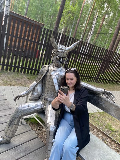 In Tal'tsy, Russia, a woman is seated on a bench next to a life-sized metal sculpture of a horned figure. The sculpture, made of welded metal plates, depicts a seated character with large horns and a stern expression, wearing a jacket and trousers. The woman is wearing a white t-shirt, a black blazer, and light blue jeans. She is holding a smartphone in her hands and appears to be taking a selfie with the sculpture. She has sunglasses pushed up on her head. The scene is set outdoors, with a wooden fence and trees in the background, suggesting a park or open-air museum. The ground is covered with wooden planks and gravel.