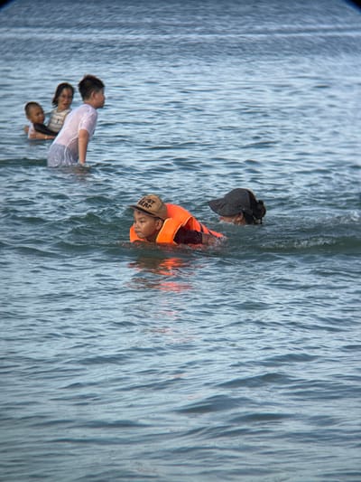 This image captures a group of people wading in the ocean near Dong Ninh Hoa, Vietnam. In the foreground, a young boy wearing an orange life vest and a baseball cap swims in the water. To his right, an adult, possibly a woman, is also in the water, wearing a dark hat and a white mask. Further back, a young boy is partially submerged, and behind him, a woman and another child are visible. The water is a moderate blue, with small waves creating ripples on the surface. The background is indistinct, suggesting open water.