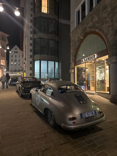 This image captures an urban street scene at night in Sankt Moritz, Switzerland. The foreground is dominated by a cobblestone or brick paved street.

A silver Porsche 356 coupe, bearing the racing number "23" on its side and "Centro Porsche Brescia" and "ENERGIAPURA" decals, is parked facing away from the viewer on the right side of the street. Its Italian license plate reads "CE 971NJ." Behind it, a dark-colored vintage convertible with its soft top down is parked, followed by a third dark vehicle further down the street.

On the immediate right, a building with a stone facade features a large arched window, which serves as a storefront for "JIL SANDER," indicated by illuminated letters above the display windows. Mannequins dressed in apparel are visible inside the brightly lit store.

To the left of the parked cars, two individuals are walking away from the camera. The person closer to the camera wears a light-colored jacket. Further down the street on the left, a traditional white building with an illuminated "Rathaus" sign and multiple flags is visible. Overhead, several streetlights illuminate the scene. A modern building with multiple glass-paned floors and visible interior lights stands between the street and the traditional building. The sky above is dark, indicating nighttime conditions.