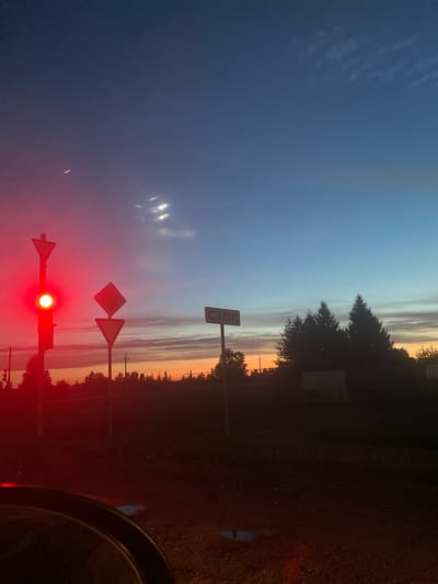 The image captures a traffic light with a red signal illuminated, set against a twilight sky displaying gradients of deep blue to orange. Several road signs are visible: a traffic light pole with a red signal and a directional arrow pointing upwards, two triangular yield signs stacked vertically below it, and a rectangular sign with Cyrillic text reading "СТОП" (STOP) to the right, mounted on a pole. The background features a silhouette of a dense line of coniferous trees and some shorter deciduous trees. A faint structure, possibly a fence or building, is discernible behind the trees on the right. The foreground shows a dark area, likely the interior of a vehicle, with a curved edge of a dashboard or steering wheel. The ground in the immediate foreground appears to be dirt or gravel with reflections, possibly from puddles. The scene is set at dusk, with ambient light originating from the sky and the illuminated traffic signal. The location is identified as Nizhnyaya Maktaama, Russia.