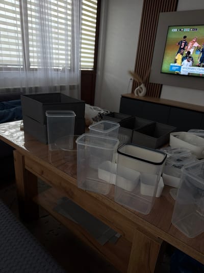 The image depicts an indoor scene, likely a living room or dining area, in an apartment in Zmeitsa, Bulgaria. The focal point is a wooden coffee table laden with various transparent plastic containers and organizers of different sizes and shapes. Some of these containers are stacked, while others are placed individually. Behind the table, a dark grey fabric storage box is visible, stacked on top of another similar box. Further in the background, a dark grey sideboard or cabinet is situated against a wall adorned with vertical wooden slats. A large television screen mounted on the wall displays a football match with players in action. To the left of the television, a minimalist white vase with a dried plant is placed on the sideboard. A window with sheer white blinds and curtains is also present, allowing natural light to filter into the room. The overall impression is one of organization or preparation for organizing household items. There are no people visible in the image.