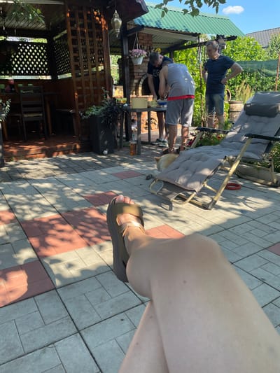 In Kobeliaky, Ukraine, on a sunny day, three people are gathered in a backyard patio area. Two men are standing, one with his hands on his hips, and another is bent over, appearing to be interacting with something on the ground. A third person's leg and foot, wearing a sandal, is prominently featured in the foreground, suggesting the photo is taken from a seated or lying-down perspective.

The setting includes a covered patio with wooden lattice work, a green corrugated roof, and a brick structure that might be an outdoor kitchen or fireplace. There are also lounge chairs, potted plants, and paving stones on the ground, indicative of a leisure space. A dog is partially visible under one of the lounge chairs.

The time of day appears to be afternoon, given the strong shadows cast by the sun. The overall mood seems relaxed and casual, consistent with a backyard gathering. No visible text is present in the image.