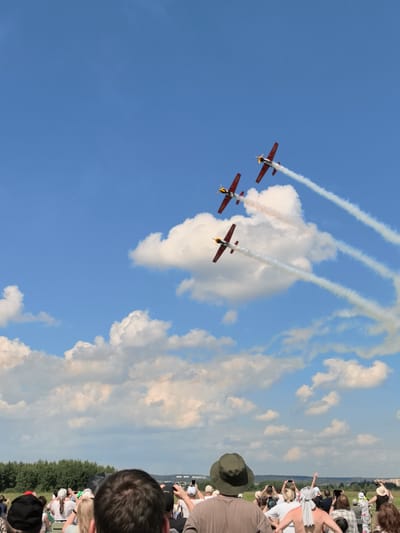 The image depicts an outdoor event, likely an airshow, with a crowd of spectators gathered in the foreground. In the sky above, three red and yellow propeller-driven aircraft are performing a synchronized flight, trailing white smoke. The sky is blue with scattered white clouds, and a line of green trees borders the horizon. The crowd consists of people of various ages, many holding up phones or cameras to capture the aerial display. The overall atmosphere appears to be one of enjoyment and observation of the performance. While the location is mentioned as Nestiukovo, Russia, this specific detail cannot be visually confirmed from the image content alone.