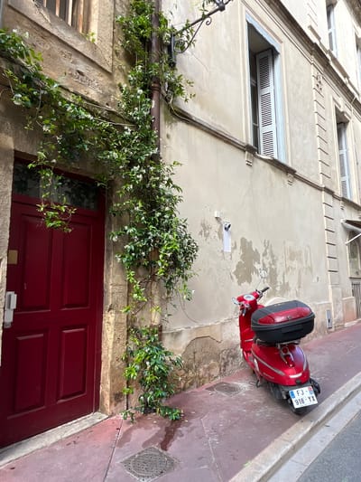 The image captures a street scene in Montpellier, France. A red scooter is parked on the side of a narrow street, adjacent to a building with a red door. The wall of the building is beige and weathered, with climbing jasmine vines cascading down the facade, adding a touch of greenery. Windows are visible in the upper portion of the building, featuring horizontal shutter designs. The sidewalk is primarily a reddish-brown color with a manhole cover. The scooter has a license plate reading "FJ-918-YX".