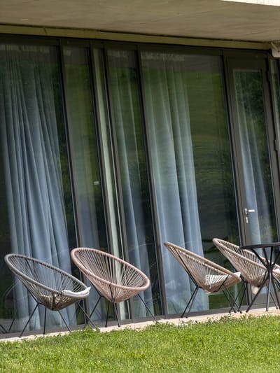 The image displays a scene with a focus on the exterior of a building. The building's facade is partially visible on the left, featuring a textured, light-colored surface. A large window with a dark frame spans a significant portion of the image, and a thin layer of window drapery are visible.

To the right of the window, there is a vertical strip of greenery, suggesting a garden or lawn. Four modern-style chairs with minimalist frames and woven seats are aligned in a row, positioned either on the grass or just beside it. The chairs are oriented at an angle, giving a dynamic composition to the shot.

The image is taken at an angle. There is no visible evidence that the image was taken in the city of Chkalov, Armenia.