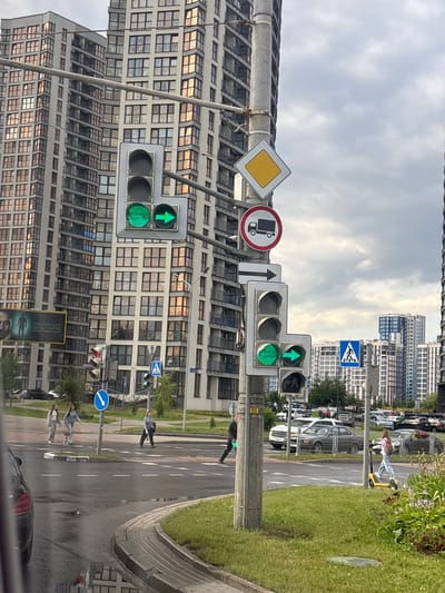 The image shows a street scene in the vicinity of Senitski Sel'ski Sovet, Belarus, with several multi-story residential buildings in the background. A traffic light pole stands prominently in the foreground, displaying multiple signals. The top traffic light shows a green circle and a green arrow pointing to the right, indicating a permissive movement for vehicles going straight and turning right. Below this, there is a red traffic light. Further down the pole, another traffic light shows a green arrow pointing to the right. Various traffic signs are also attached to the pole, including a diamond-shaped yield sign, a circular sign with a truck symbol crossed out (prohibiting trucks), and a rectangular sign with a rightward arrow.
In the middle ground, a pedestrian crossing is visible with several people crossing the street. Some individuals are walking, and one person is riding a yellow electric scooter. Cars are present on the road, and a parking lot filled with vehicles is visible in the distance. The ground is wet, suggesting recent rainfall, with puddles reflecting the surroundings. The sky is overcast with clouds. A billboard advertising "АЛЕССАНДРО ДЕЛЬ ПЬЕРО" is partially visible on the left side of the image.