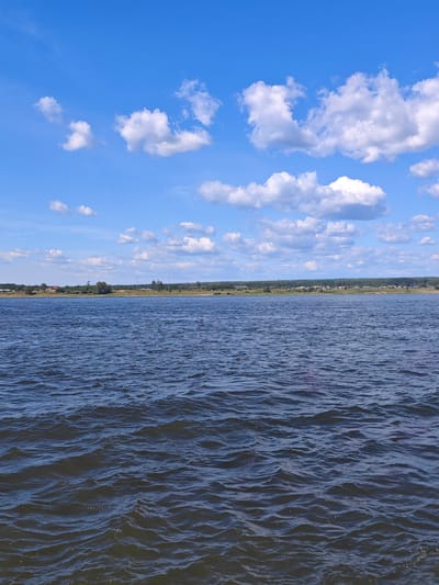 The image displays a serene body of water, likely a river or lake, under a bright blue sky dotted with fluffy white clouds. The water's surface is gently rippled, reflecting the sunlight. In the distance, a tree-lined shore is visible, with hints of buildings and structures suggesting a settlement or town. The overall impression is one of natural tranquility and a peaceful rural or semi-rural setting. The location is identified as Vysokogorskii, Russia, which is a district in the Republic of Tatarstan, Russia.