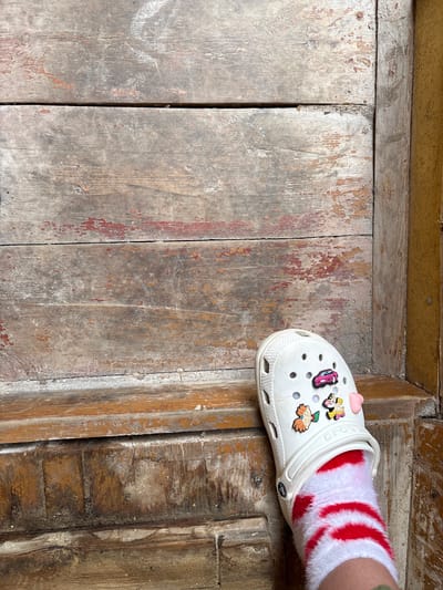 A white Croc shoe with various colorful Jibbitz charms, including a pink car, a character with blonde hair, and a pink heart, is worn by a person with fuzzy red and white striped socks. The shoe and sock are positioned on weathered wooden steps that appear to be located on the exterior of a building. The wood is rough, with peeling paint in shades of red and brown, and there are visible gaps between the planks. The background consists of more of this aged wooden structure, with a yellow-painted wall visible on the right side of the frame. The setting suggests an outdoor environment, possibly in a residential or public area with a rustic aesthetic. The specific location of Zheleznodorozhnyi, Russia, is inferred to be the context of this scene.