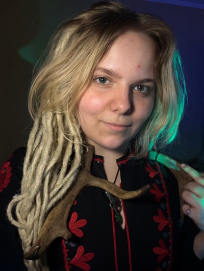 The image is an upward-facing, close-up, studio portrait of a young person. They have light blonde hair with streaks of teal at the ends, and their hair is styled in loose waves. The person's eyes are a light green-grey, and they have a light dusting of freckles across their nose and cheeks. They are wearing a black garment with red floral embroidery. The person is holding a deer antler, which is positioned across their chest and extends out of frame. The background is dark with a blue and green gradient, suggesting artificial lighting. The overall mood is serene and somewhat mysterious. There is a ring with a purple stone on the person's right ring finger. A silver pendant with a green stone hangs on a red cord around their neck. The image appears to have been taken indoors, likely in a home studio. The Vostochny District, Russia is mentioned as a location context, but there are no specific visual cues in the image to confirm this.