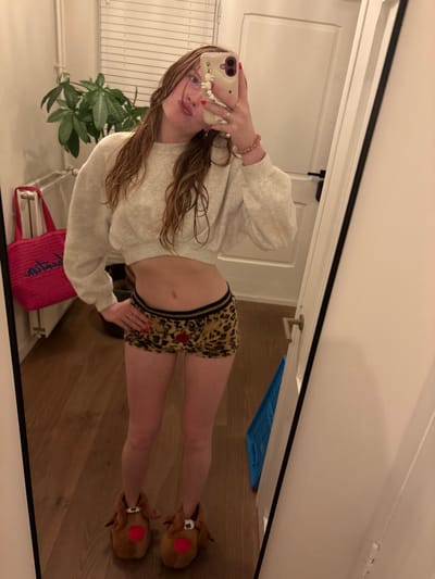 A woman takes a mirror selfie. She wears a light-colored sweater, leopard-print shorts with a red heart, and reindeer slippers. She holds a phone with a cream-colored case and a white charm attached. The background includes a white door, a small houseplant, a pink tote bag, and a wooden floor. The image is rotated, with the top of the frame on the right side.