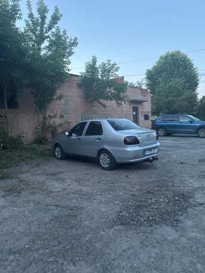 An outdoor, ground-level, eye-level medium shot captures a silver Fiat sedan parked on a gravel lot in front of a brick building in the city of Гожули, Ukraine. The building, partially obscured by trees, appears aged and has some graffiti on its walls. To the right of the Fiat, a blue SUV is also parked. The sky above is a clear blue, with a few dark objects that appear to be birds or insects. The scene is quiet and still, with no people present. The lighting suggests it is either early morning or late afternoon. The Fiat has a Ukrainian flag decal on its rear, and its license plate reads "BI 9210 IM".