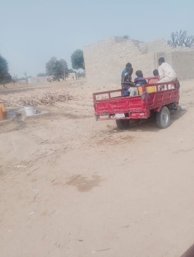 The image depicts a rural or semi-urban scene in Kanwa, Nigeria, under clear, bright daylight. The foreground consists of light brown, dry, dusty earth with some faint tire tracks and scattered debris.
In the midground, a red three-wheeled motorized vehicle, appearing to be a cargo tricycle with a bed, is positioned slightly off-center to the right. Four individuals, all male, are seated or standing in the cargo bed. One man, wearing a light-colored traditional garment (agbada or similar), stands at the rear of the bed, facing forward. To his left, another man in a dark shirt with light stripes sits. Next to him, a younger individual in a dark blue shirt is seated. A fourth individual, partially obscured, is also visible. Two yellow plastic jerrycans or containers are visible within the cargo bed, near the seated individuals. The vehicle's single rear wheel and a license plate are visible.
To the left of the vehicle, on the dusty ground, there is a pile of dry, light-colored wooden sticks or branches, possibly firewood or construction material. A white bucket and a smaller yellow object are near this pile.
The background features several structures constructed from light-colored concrete blocks, appearing to be partially built or in disrepair, suggesting a construction site or an undeveloped area. Several sparse trees with green foliage are scattered across the horizon, with the sky being a pale blue, almost white, indicating either haze or bright sunlight.