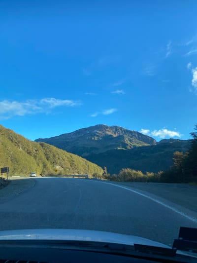 The image depicts a winding asphalt road through a mountainous landscape near Lago Escondido, Argentina, observed from the interior of a vehicle. The sky is a clear, bright blue with sparse, wispy white clouds across the upper half of the frame. In the mid-ground, a prominent mountain range rises, featuring rugged, dark green and brown forested slopes, with patches of snow or ice visible near the peaks.
The two-lane road curves leftward into the distance. A solid white line marks the right edge of the visible pavement, and a broken white line indicates the center or passing lane on the left side of the curve. A white passenger vehicle, possibly an SUV, is visible ahead on the left side of the road, traveling in the same direction as the camera. Roadside elements on the left include a dark rectangular sign, a yellow diamond-shaped warning sign indicating a curve, and a white guardrail. Dense vegetation, primarily trees with varying shades of green and some brown, covers the slopes adjacent to the road. The immediate foreground shows the dashboard and a portion of the windshield of the vehicle from which the photograph was taken. The lighting suggests bright daylight.