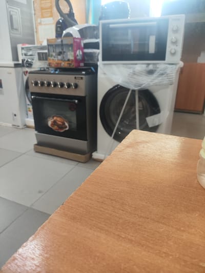 The image displays a collection of household appliances in what appears to be a store or showroom, possibly in Kerben, Kyrgyzstan. In the foreground, a wooden surface dominates the lower portion of the frame. Behind this, a stainless steel gas oven with a glass door is prominently featured. The oven displays a circular image of roasted chicken with vegetables inside its oven cavity. Above the oven, a white microwave oven is visible, with its door slightly ajar. To the right of the oven, a white washing machine is partially obscured by plastic packaging. Further back, the scene includes other appliances, including what looks like a refrigerator on the left, and the silhouette of a vacuum cleaner. The setting is brightly lit, suggesting an indoor environment. The overall impression is of a retail space showcasing kitchen and laundry appliances. There is no discernible activity or event taking place, as the appliances are stationary and there are no people visible. The time of day and weather are not determinable from the image.