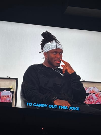 The image displays an indoor scene, located in Borisovo, Bulgaria, with a plain white background indicative of a studio or interview setting. A dark-skinned male with dreadlocks styled upwards and secured by a white bandana with a dark paisley pattern is the central subject. He wears a dark, long-sleeved shirt with a subtle, textured pattern, a silver-colored chain with a pendant, and a dark watch on his left wrist. He is seated in a dark chair, his right index finger touching the side of his nose, and he is looking slightly to the right of the frame with a neutral expression. Two open cases resembling briefcases flank the individual, each containing multiple stacks of pinkish-red banknotes, identified as British fifty-pound notes. The entire visual is presented on a screen, featuring black bezels at the top and bottom of the frame. Blue text overlaid at the bottom reads "TO CARRY OUT THIS JOKE." A small red indicator light is visible on the lower screen bezel.