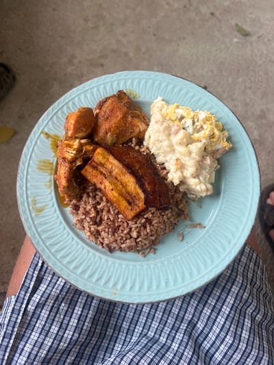 The image shows a plate of food, likely being held by someone. The plate contains rice and beans, fried plantains, a serving of what appears to be potato salad, and pieces of chicken. The person holding the plate is wearing a blue and white checkered shirt or pants. The background includes a glimpse of what looks like concrete flooring and part of a shoe, suggesting the scene is likely outdoors or in a casual setting. There is nothing to indicate the specific location of Punta Gorda Town, Belize, beyond the likely regional cuisine on the plate.