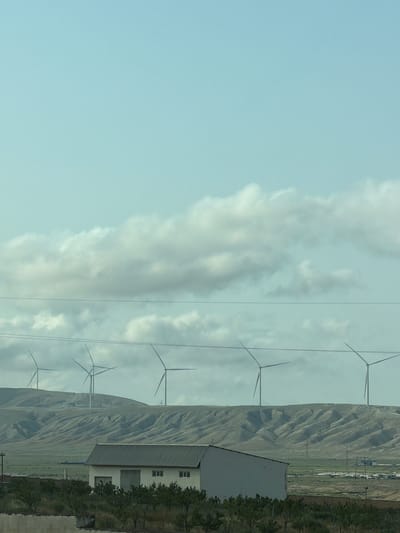In Pirəkəşkül Qəsəbəsi, Azerbaijan, a wind farm is visible on a distant hillside. Several wind turbines are spaced out across the undulating terrain. In the foreground, a long, low building with a sloped roof and several windows stands. The sky above is a pale blue with scattered clouds. The landscape appears arid and hilly, with sparse vegetation in the foreground.