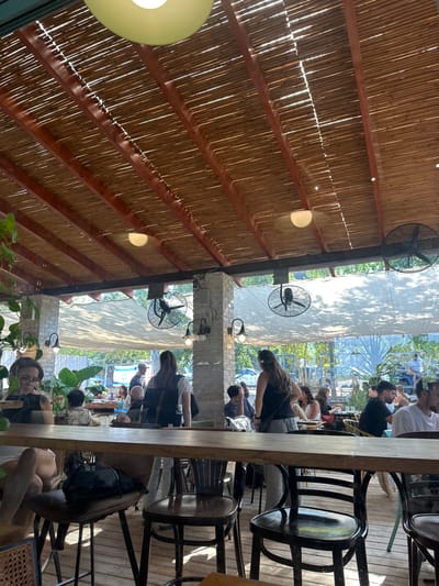 The image depicts a semi-outdoor restaurant or cafe located in Kfar Monash, Israel, observed from a low-angle perspective, looking towards the back of the establishment. The environment is brightly lit by natural sunlight filtering through a slatted wooden and bamboo-style ceiling structure, supported by light-colored stone or brick pillars. Recessed dome lights and hanging pendant lights are integrated into the ceiling, along with several industrial-style wall-mounted fans attached to the pillars and beams, circulating air.

In the immediate foreground, a long, light-colored wooden bar-height table extends across the frame. To the left, a person with glasses is partially visible, seated at this table, looking downwards at an unseen object. Their right leg and foot are visible under the table, and a dark bag rests on a stool beside them.

The midground features numerous individuals. To the left, a woman wearing a light-colored top and a dark backpack stands with her back mostly to the camera, facing right. Near the center, another woman with long dark hair, wearing a dark top, stands with her back partially to the camera. Groups of people are seated at various dark wooden tables and chairs throughout the space, engaged in conversation or dining. A small child is visible in a high chair at a table further back.

Beyond the main seating area, a large white fabric awning or shade sail is visible, casting dappled shadows. More people are seated at tables in this area. In the far background, a dense line of trees suggests a boundary, and a person in a light blue shirt is seated on a low wall or bench. The flooring appears to be made of wooden planks. The overall scene indicates a busy, casual daytime setting.