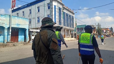 The image depicts an outdoor urban street scene in Gisenyi, Rwanda, during clear daylight. Four individuals, identified as street clean-up personnel by their reflective vests and brooms, are observed walking away from the camera down a paved road.

In the immediate foreground-left, an individual wears a camouflage bucket hat and a dark olive-green jacket with a brown patch on the upper right arm, carrying a long stick-like tool. To their right, a second person wears a dark cap, a yellow-green reflective vest over a plaid shirt, and white gloves, holding a broom. Centrally positioned, a third person wears a dark blue cap, a dark blue shirt, and a yellow-green and blue reflective vest with white text "MAIRIE DE GISENYI" and additional Kinyarwanda script, wearing orange gloves and holding a broom handle. Further down the street on the right, a fourth individual in a yellow-green reflective vest and white footwear holds another broom.

The street is asphalt. On the left, a light blue two-story building with a reddish-brown double door and steps is visible, with a red pole displaying partial text "BROS" and "QUALITES." Behind it stands a multi-story light gray building with dark blue-framed windows. On the right, a modern multi-story building features a light gray facade and blue reflective glass panels. Various other commercial and residential structures line the street into the distance. Numerous overhead utility lines traverse the light blue sky, which contains scattered white clouds.