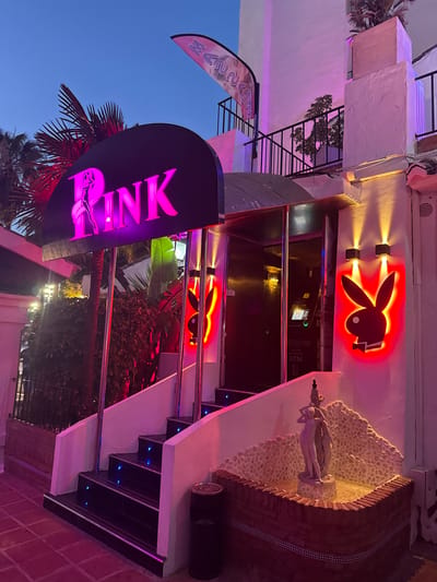 The image shows the exterior of a building at dusk, illuminated by neon lights. A prominent sign with the word "PINK" in large, glowing pink letters is visible, featuring a stylized silhouette of a woman. Above the entrance, a red neon Playboy bunny logo is displayed.
The building appears to be a commercial establishment, possibly a nightclub or bar, with an entrance featuring a set of stairs. The stairs are lit with small blue lights integrated into the risers. To the right of the entrance, a small fountain with a statue of a reclining nude figure is set into a wall adorned with pebbles and brick. Palm trees and other foliage are visible around the building, suggesting a warm climate. The sky is a deep blue, indicating twilight.
A banner with stylized lettering that reads "MANSA" is also visible on the left side of the image. The overall mood is vibrant and alluring, consistent with nightlife establishments. The location cues, such as palm trees and the style of architecture, combined with the given location context of Nueva Andalucía, Spain, suggest this is a venue in a popular tourist or entertainment area. There are no people visible in the image.