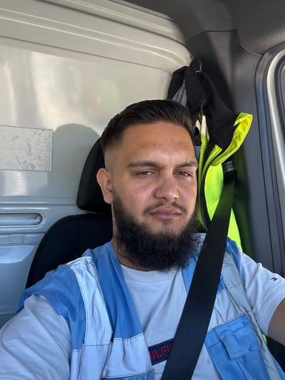 The media file is a close-up, sideways shot of a man in a vehicle. The man has dark hair, a beard, and brown eyes. He is wearing a white t-shirt with blue accents, a high-visibility yellow and black vest, and a black seatbelt across his chest. The man appears to be sitting in the passenger seat of a truck or van, as indicated by the interior paneling and the seatbelt. The background shows the interior of the vehicle, including a black headrest and parts of the dashboard. The lighting suggests it is daytime, but the weather is not discernible. The man's expression is neutral, with a slight smile. The overall setting appears to be casual, possibly during a commute or a break. There is no discernible text in the image. The location context provided is Stavelot, Belgium, which is not visually confirmed by the image itself.