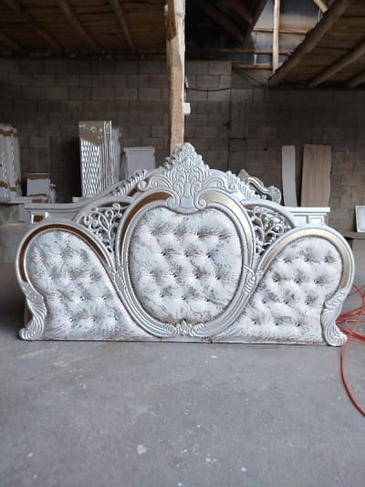 A highly ornate, tufted headboard, predominantly white with silver-grey and gold accents, rests on a light grey concrete floor within a furniture workshop or storage area in Andijan, Uzbekistan. The headboard features a central oval-shaped upholstered panel flanked by two smaller, curved, rectangular upholstered panels, all deeply tufted with decorative crystal-like buttons. Its carved wooden frame displays elaborate scrollwork and is finished in a metallic silver-white with gold trim around the upholstered sections. In the background, a light grey cinder block wall is visible. To the left, several white or light-colored furniture components, including a tall, vertically fluted piece, are stacked or arranged. To the right, two light-colored wood panels lean against the wall, and a coil of red electrical wire lies on the concrete floor. Exposed, rough-hewn wooden beams form the ceiling structure above. No individuals or explicit actions are depicted.